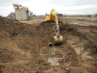Excavation of soil for pipe inspection, with one of several former ammunition plant structures in background